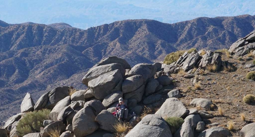 A person sits near a rock formation. There are mountains in the background. 